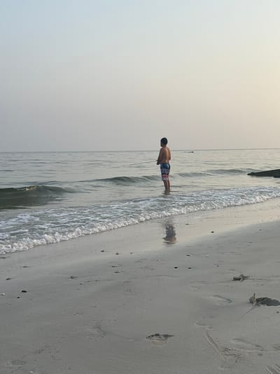 An individual stands in the shallow ocean water at a beach, holding a fishing rod and casting a line. The person is wearing multi-colored swim trunks and their back is to the camera. The water is calm with gentle waves lapping at the shore. The sand is damp and shows footprints from previous activity. The sky is overcast and hazy, suggesting early morning or late afternoon light. The scene is likely a beach in Ajman, United Arab Emirates, given the style of the swim trunks and the general appearance of the beach and water.