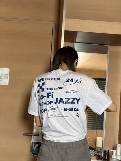 A person with short dark hair, wearing a white t-shirt with music-themed graphics in dark blue, is leaning over a bathroom sink. The text on the t-shirt includes "WE LISTEN," "THE MUSIC," "Lo-Fi," "HIPHOP JAZZY," "POP & B-SIDE," and "2413." A short phrase about refuge is also printed. The person is wearing grey sweatpants. In the background are a mirror, a sink with faucet, and a light brown wall and door. A sign written in Japanese indicating the room is occupied is on the door handle.