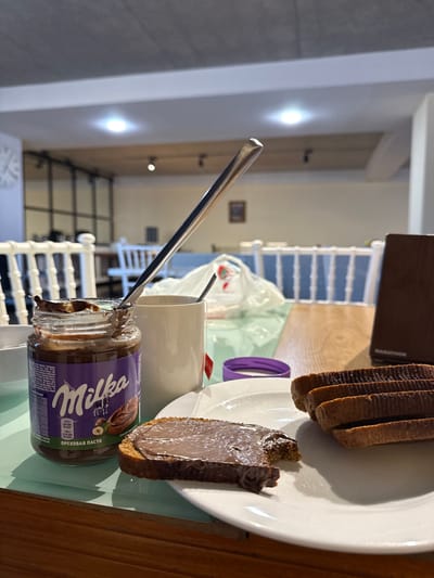 The image displays a close-up, slightly elevated view of a breakfast or snack scene indoors, located in Bakuriani, Georgia. In the immediate foreground, on a light green transparent mat covering a wooden table, a white ceramic plate holds a partially consumed slice of dark bread spread with a brown chocolate-hazelnut paste. Adjacent to this, three unspread slices of identical dark bread are stacked on the same plate. To the left of the plate, an open glass jar of Milka hazelnut spread (labeled "ОРЕХОВАЯ ПАСТА") contains a spoon. Behind this jar, a white ceramic mug has a red tea bag tag visible, with a second spoon inside the mug. A purple plastic lid is partially visible behind the mug. A long, slender silver spoon leans diagonally, extending from the Milka jar upwards and to the right. The background is an out-of-focus interior space, characterized by multiple white chairs with spindle-back designs, some featuring blue seat cushions, indicative of a communal dining area or cafe. The ceiling is grey, revealing exposed wooden beams and recessed lighting fixtures. A portion of a white-faced wall clock is visible on the far left, and a framed picture hangs on a distant wall.