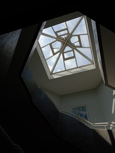 The image shows a view looking up towards a geometric skylight with a view of the sky and clouds. Below the skylight, a staircase with a railing is visible, curving upwards. A painting is hung on the wall next to the staircase. The architecture appears modern, with sharp angles and a mix of dark and light surfaces. The lighting suggests it is daytime, with sunlight filtering through the skylight and casting shadows. This scene is located in the main hall of the University of Constance, Germany, a building known for its distinctive architecture.