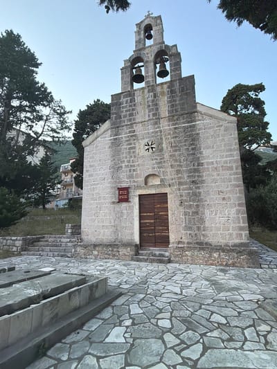 The image shows a stone church with a bell tower, located in Medinski Krš, Montenegro. The church has a wooden double door and a small window with a decorative element above it. The bell tower has three bells and a cross at the top. The ground in front of the church is paved with irregular stone tiles. There are some stone structures in the foreground that appear to be tombstones. Trees surround the church, and a multi-story building is visible in the background, suggesting an urban or semi-urban setting. The sky is clear and light blue, indicating daytime.