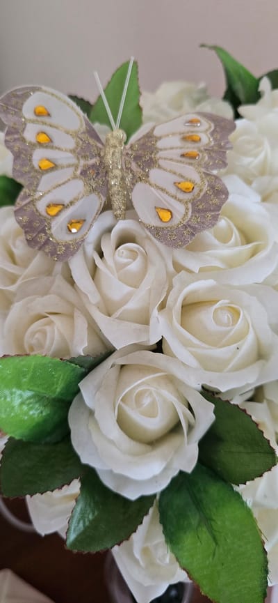 A close-up, slightly angled shot shows a decorative artificial butterfly perched on a bouquet of white roses. The butterfly is predominantly white and light purple, with gold glitter accents and small, orange jewel-like embellishments on its wings. Its body is golden and glittery, with two thin, white antennae extending upwards. The white roses are densely packed, with visible green leaves scattered throughout. The background is softly blurred, suggesting an indoor setting with a neutral-colored wall. There are no people or discernible actions depicted in the image. The overall impression is of a decorative arrangement, possibly for an event or as a gift. The specific location of Dzhalka, Russia, is not indicated by any visual cues in the image.