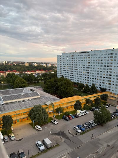 An elevated, eye-level, wide-angle exterior shot captures a residential district in Solna, Sweden, at dusk. The sky above is a canvas of muted grey and pink clouds, hinting at the setting sun. Dominating the right side of the frame is a massive, light blue, multi-story apartment building with a grid of numerous windows, some illuminated from within, suggesting occupied residences. In the middle ground, a long, low-rise building with a distinctive orange-yellow covered walkway stretches horizontally. This building appears to be a school or community center, with "SUNNAN" visible on its exterior. Adjacent to this, a parking lot is filled with a variety of cars, including sedans, SUVs, and vans, parked in designated spots. Beyond these structures, a dense cluster of residential buildings with red-tiled roofs and green trees forms a suburban landscape. Further in the distance, more buildings and trees suggest the expanse of the city. A single person is visible in the lower right of the frame, walking along the paved road. The overall impression is one of a typical urban evening scene in a residential area.