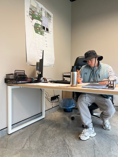 The image depicts a person seated at a desk in an office setting. The person is wearing a hat, a light-colored vest, and grey pants. They are sitting on a rolling chair, with their legs extended in front of them. On the desk, there is a computer monitor, a water bottle, paperwork in a clip, and other office supplies. A large blueprint is visible on the wall. The floor is tiled, and the walls are painted in a neutral color. Based on the Зеленогорское сельское поселение location, the image was taken in Ukraine.