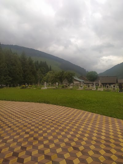 A cemetery is visible in the foothills of Luhy, Ukraine, with a cloudy sky overhead. The cemetery is filled with numerous gravestones and crosses of various sizes and designs, interspersed with green grass and trees. In the background, the landscape rises into forested hills. In the foreground, a patterned walkway made of brown and yellow interlocking bricks leads the eye into the scene. Several rustic buildings with wooden roofs are situated behind the cemetery.