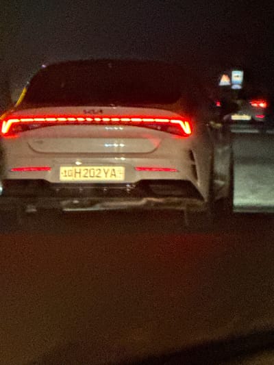 A close-up, grainy, and tilted image depicts the rear of a white Kia EV9 at night. The car's vertical LED taillight is prominently illuminated, as is the license plate, which reads "10 H 202 YA". The license plate has a yellow background. Part of a large vehicle is seen above the car. The surrounding environment appears dark, with a muted brownish area on the right side.
