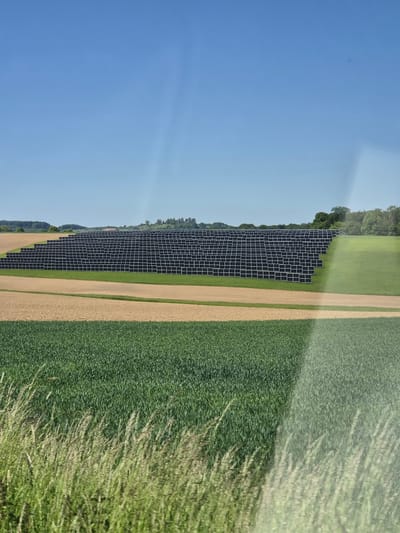The image depicts a solar panel array located near Lenningen, Luxembourg. The panels are situated between a dirt or gravel road and a field of green crops. A line of trees borders the panels on one side, transitioning into an open field. The sky is clear and blue. The image appears to be taken from a moving vehicle, possibly through a window, as evidenced by the blurry reflections.