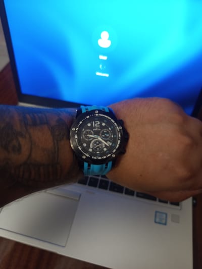 A close-up, overhead shot shows a person's left arm and wrist wearing a black and blue Invicta watch. The arm has a tattoo on it. The watch has a black face with blue accents and a tachymeter bezel. The watch is positioned over a laptop keyboard. The laptop screen is visible in the background and displays a blue login screen with a user icon. The scene appears to be indoors, likely in a home or office setting, with no indication of the city of Radishevo, Bulgaria.