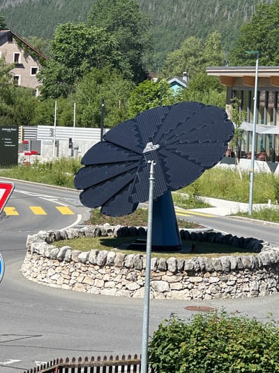 In Le Brassus, Switzerland, a unique solar-powered flower-shaped sculpture is centrally located in a traffic circle. The sculpture, composed of dark solar panels arranged like petals, stands on a blue cylindrical base. It is surrounded by a circular stone wall, with grass and small plants within. A grey metal pole, likely a light post or part of the sculpture's mechanism, is positioned in front of it. A traffic sign with red and white diagonal stripes is visible on the left, and yellow pedestrian crossing markings are painted on the asphalt road encircling the sculpture. In the background, several buildings are visible, including a beige structure with red-trimmed windows and a modern building with large windows and outdoor seating areas. Lush green trees and a forested hillside form the backdrop. The scene is captured on a sunny day.