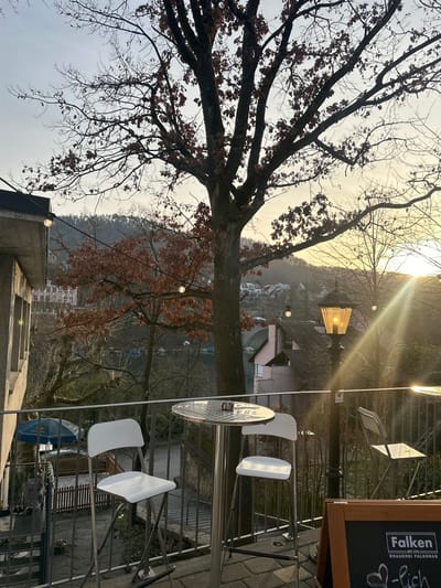 The image displays an outdoor terrace located in Eglisau, Switzerland, overlooking a river and a hilly, residential landscape. The scene is illuminated by late afternoon or early evening light, characterized by sun rays emanating from the right side of the frame.
In the immediate foreground, a paved terrace surface features two round, silver-colored high-top tables, each accompanied by two white bar stools with chrome frames. An ashtray is present on the left table. To the far right, a partial view of a black chalkboard sign displays the text "Falken Brauerei Falkenag." String lights with spherical bulbs are suspended overhead, some of which are illuminated. A black lamppost, centrally positioned near the right table, features an illuminated lantern casting a warm light and creating a sun flare effect. A metal railing delineates the edge of the terrace.
Beyond the railing, a descending path or staircase is visible. A large deciduous tree, mostly bare with some retained brown leaves, stands prominently in the midground, partially obscuring the view. Further beyond, a greenish-blue river flows, bordered by various residential structures, including a distinctive pinkish building and a stone arch bridge in the distance. The background consists of wooded hills covered with mostly bare trees, set against a pale, bright sky where the sun's position suggests either sunrise or sunset. No individuals are visible anywhere in the scene.