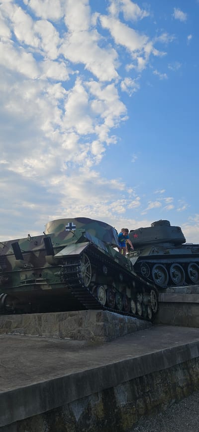 In Svidník, Slovakia, two historical tanks are displayed on a concrete and stone embankment. The tank in the foreground is painted with a camouflage pattern and features a white cross insignia on its turret. A young boy in a blue t-shirt and shorts is climbing on top of this tank, reaching towards the second tank behind it. The second tank is dark green and has a red star insignia on its turret. The sky above is a clear blue with scattered white clouds. The scene appears to be outdoors, possibly in a park or memorial site.