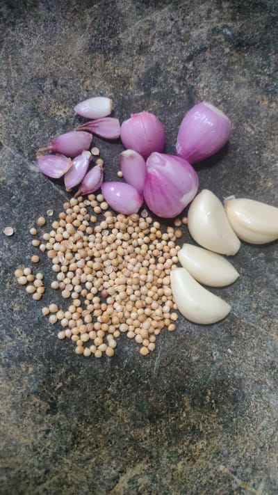 The image shows a collection of ingredients for cooking, likely in a kitchen or outdoor cooking space in Bojong Gede, Indonesia, located at the provided coordinates. The ingredients are arranged on a rough, circular, dark gray, speckled surface, possibly a mortar and pestle or a stone slab. The ingredients include:
* a large pile of coriander seeds in the center
* six red onions, some whole and some halved, arranged around the coriander
* three cloves of garlic, halved, in a semi-circle on the right side of the coriander
* one small piece of red onion skin
* several individual coriander seeds scattered nearby
* several small pieces of red onion skin.
The ingredients are arranged in a visually pleasing manner, suggesting that they are ready for use in a dish. The overall scene is one of preparation, with a sense of anticipation for the cooking process to come.