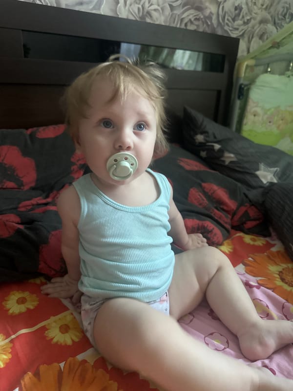 Infant resting on a bed in Sayanogorsk, bottle and pacifier nearby.