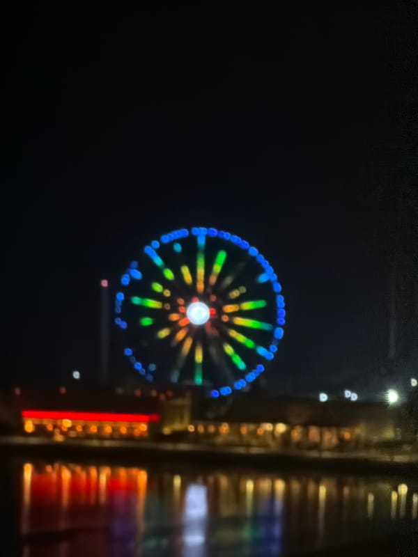 Lit-up Ferris wheel at night, viewed over water with coffee in hand