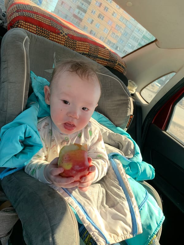 Baby in dinosaur pajamas enjoys apple snack in Izhevsk car seat