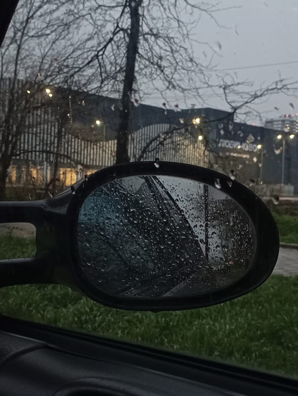 Rain-covered car mirror reflects blurred Krasnodar street scene