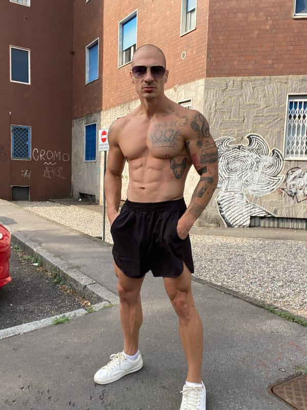 Tattooed figure poses outside brick apartment building in Corsico