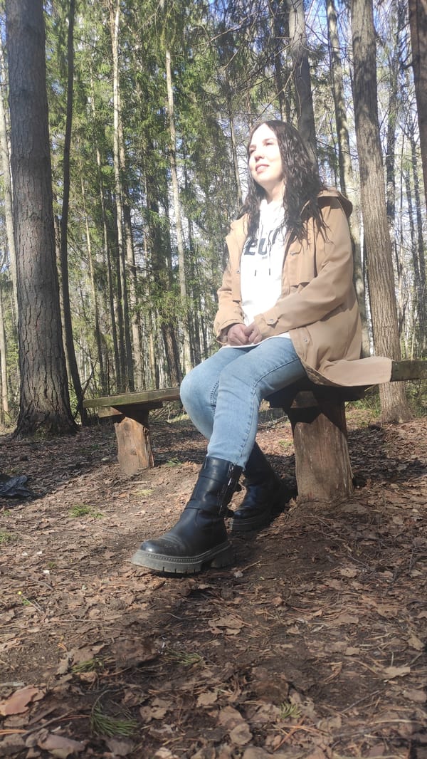 Woman in tan jacket sits on wooden bench in forest