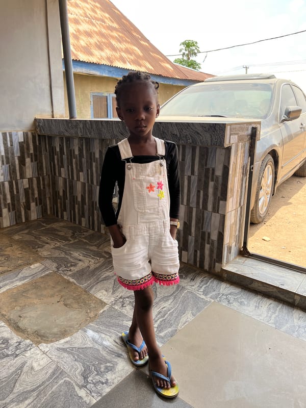 Girl in floral overalls stands outside building in Madalla