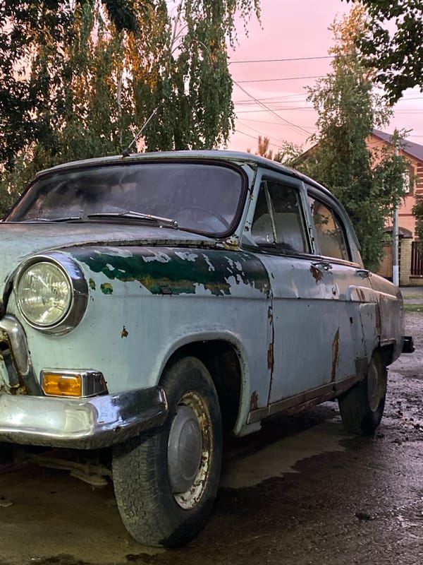 Rusted blue sedan parked on wet asphalt in Dinskaya