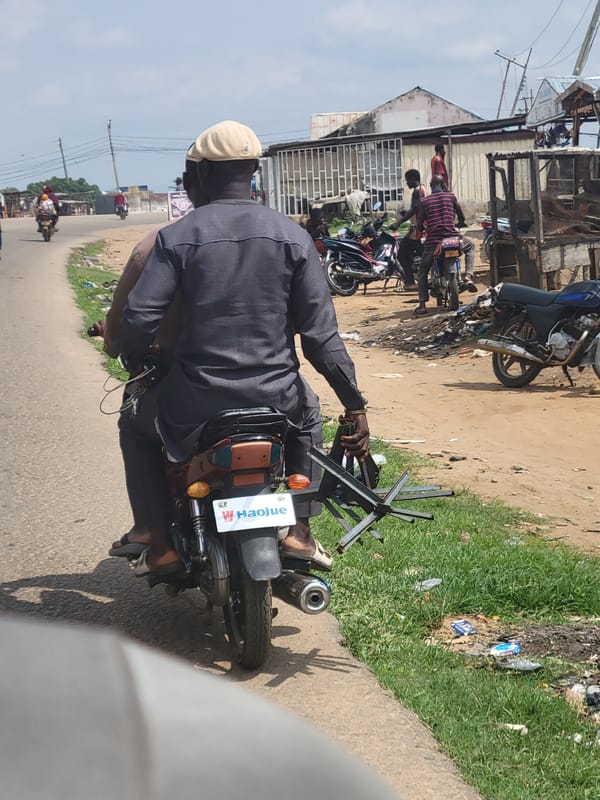 Two men ride motorcycle on dirt road in Keffi