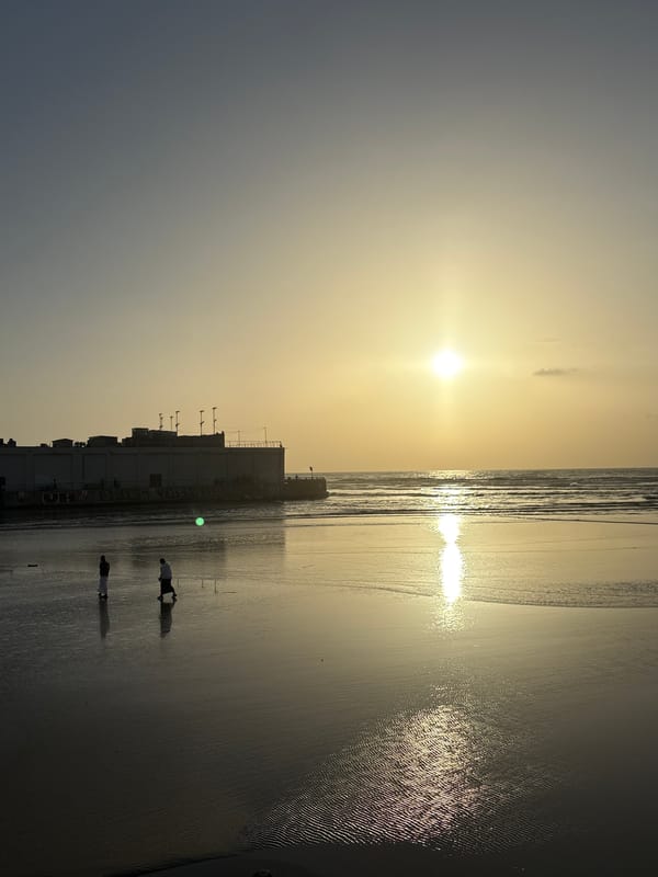 Two people walk on beach at sunset near shoreline building