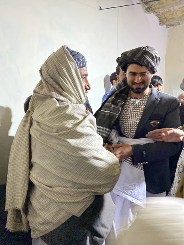 Men gather indoors wearing traditional attire in community meeting