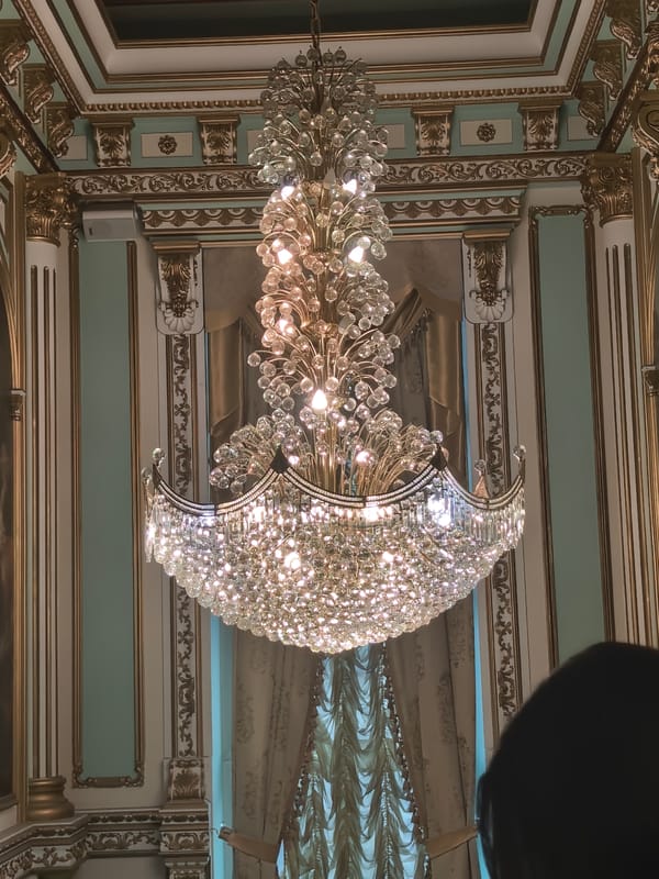 Inside a gilded ballroom or palace, chandeliers and classical murals throughout
