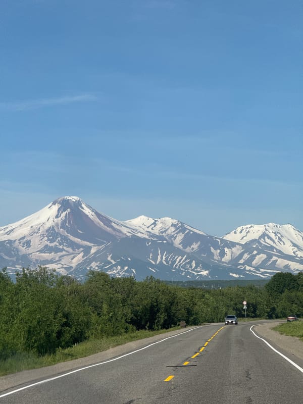 Snow-capped volcanoes loom over a curving road in Kamchatka
