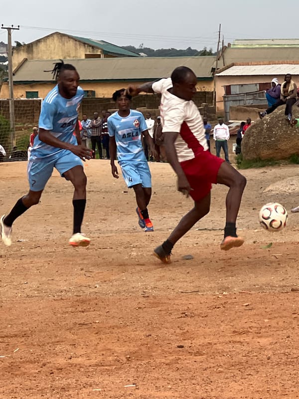 Football on a dirt pitch in Jos, Nigeria, rocky plateau backdrop.