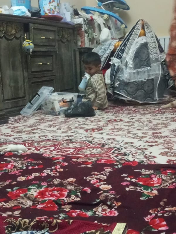Child drinks from bottle on rug near household items