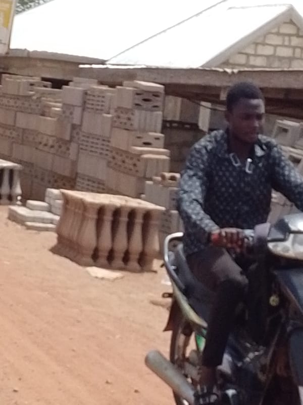 Motorcycle rider travels dirt road past construction materials in Dungu