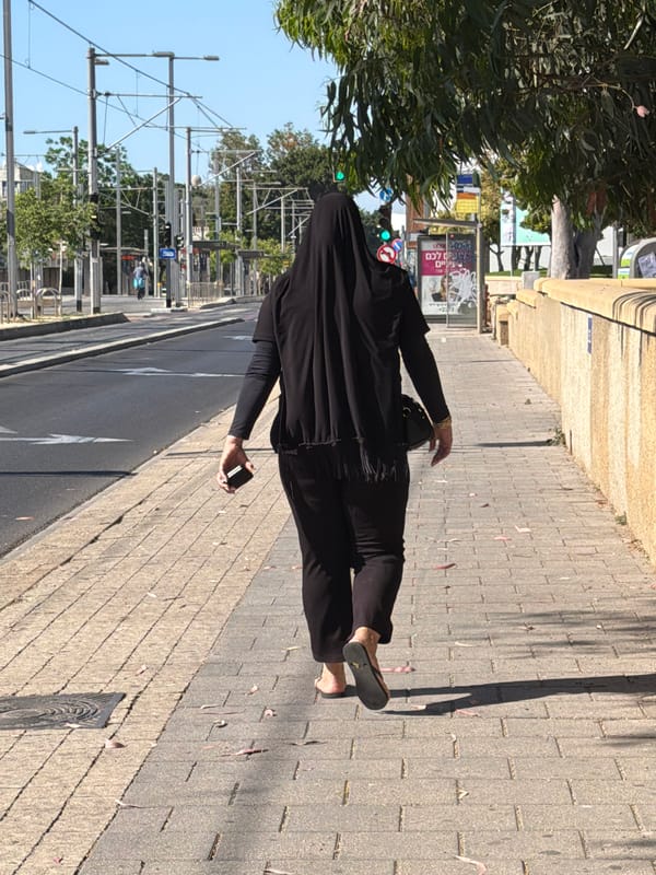 Person in black walks sidewalk with phone in Bat Yam