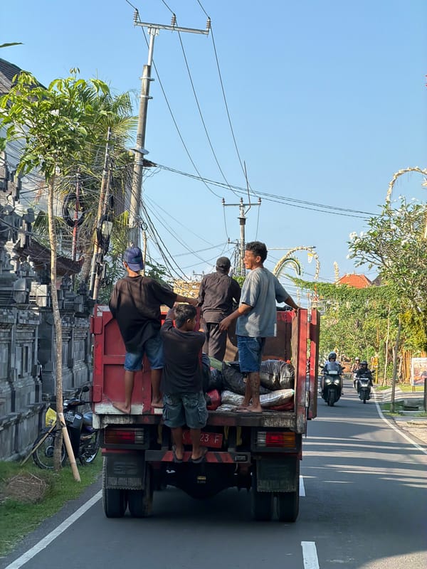 Red flatbed truck carries passengers through sunny Mengwi streets
