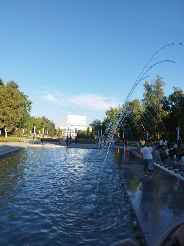 Summer afternoon at a Shymkent park — fountains, play structures, crows.