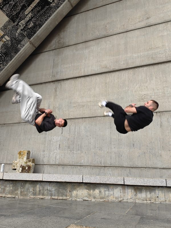 Two men perform synchronized aerial acrobatics near concrete structure