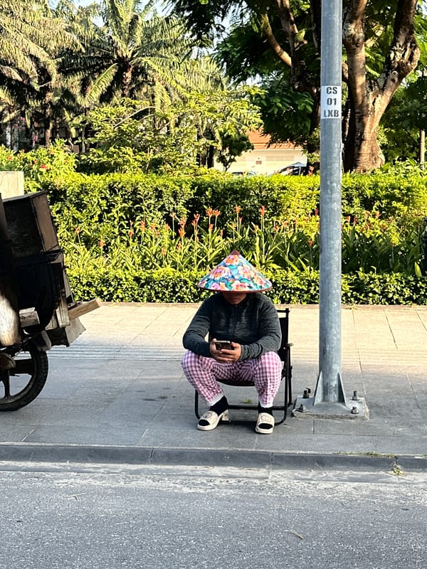 Elderly man in traditional hat uses phone on Đà Nẵng sidewalk