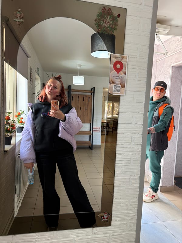 Woman takes mirror selfie with man in background, Noviy
