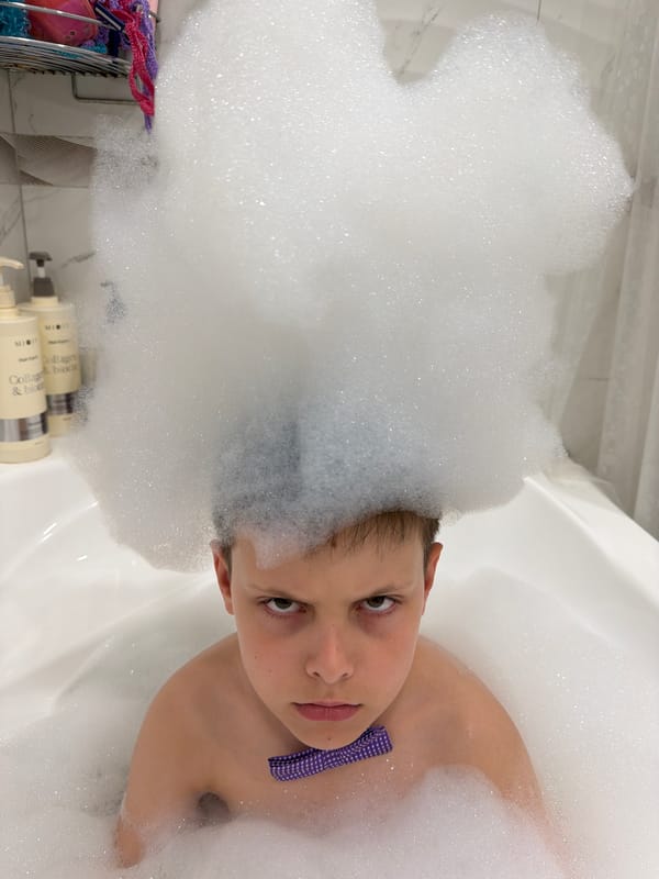Child takes bubble bath wearing glasses and bow tie