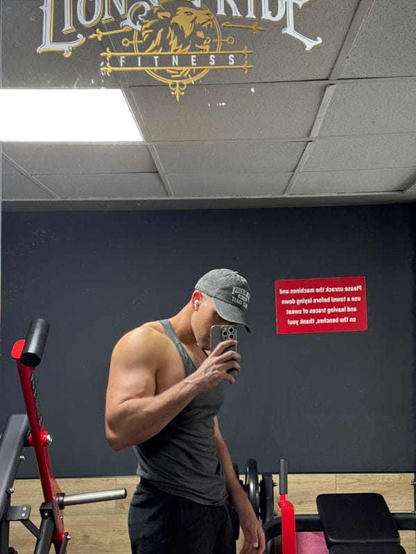 Man takes mirror selfie at gym wearing grey tank