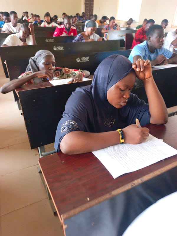 Students writing at desks in classroom, hijab-wearing student visible