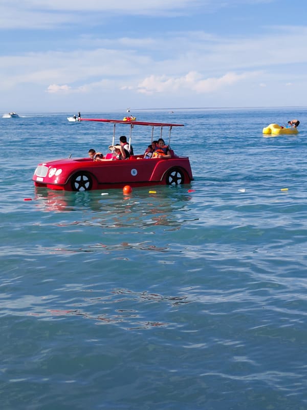 Crowded pebble beach day in Ақші with pedal boats and swimmers