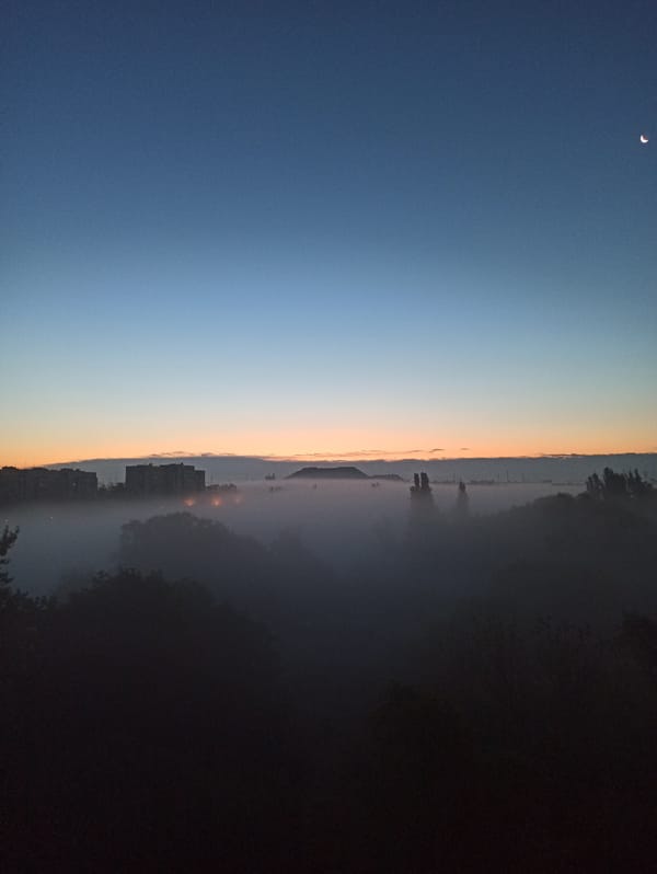 Crescent moon over a fog-wrapped industrial skyline at dusk