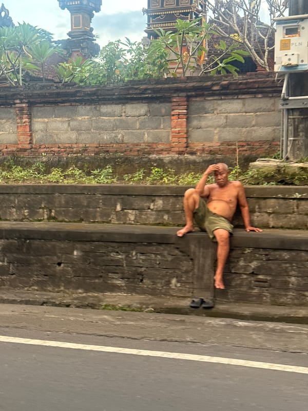 Elderly man rests on roadside wall in Sukawati