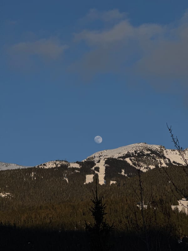 Moon visible over snow-covered Whistler mountains under blue sky