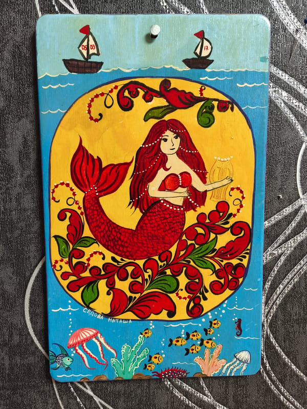 Colorful folk art paintings observed featuring mermaids and village scenes