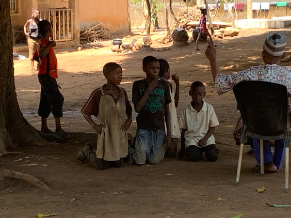 Four boys kneel in village setting with adult present