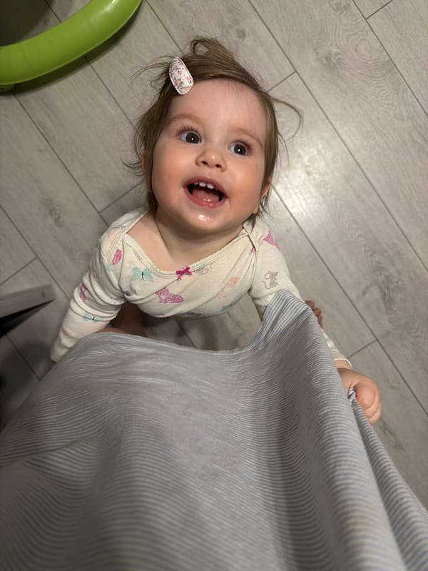 Child in onesie holds curtain on wooden floor