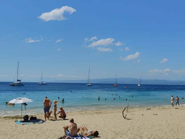Busy Greek beach morning — swimmers, cafe, sailboats, coastal flora