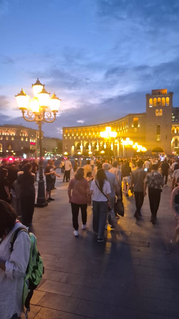 City square at dusk, crowds moving under glowing streetlamps